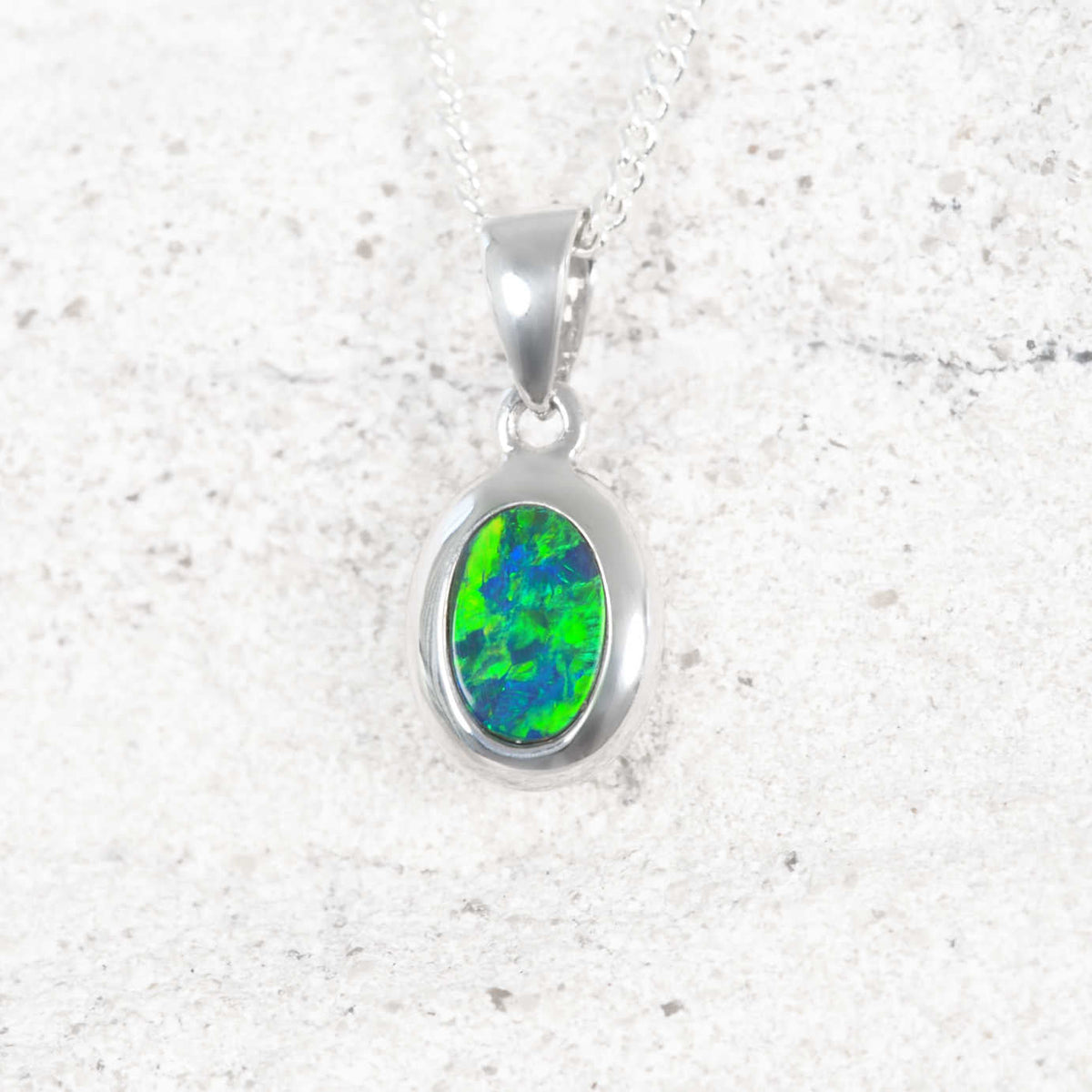 A white opal authentic oval opal pendant necklace featuring a vibrant green and blue opal stone, displayed on a textured light gray stone surface.