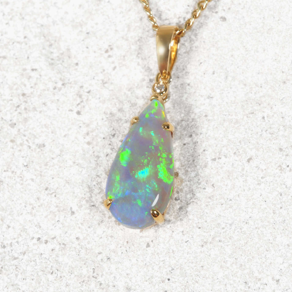A teardrop-shaped authentic Australian opal pendant necklace in a gold setting on a textured light gray background. The opal displays vibrant green, blue, and white flashes, set in a gold bail with a small clear accent stone.