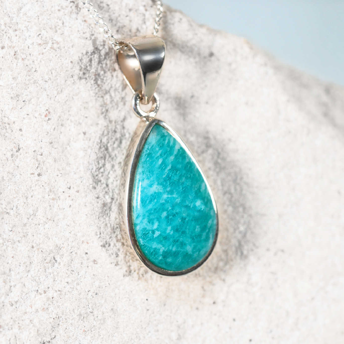 Teardrop-shaped turquoise pendant on a silver chain against a sandy background