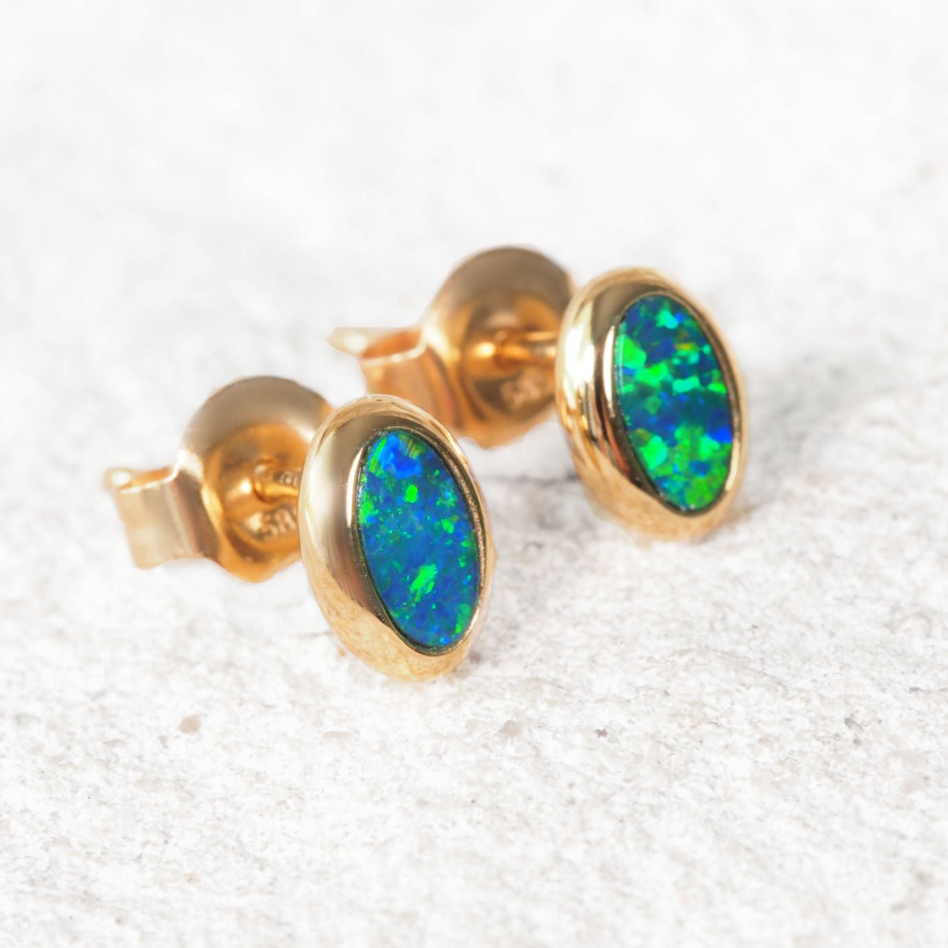 A pair of gold oval Australian opal stud earrings with vibrant green and blue opal inlays, shown on a textured white background.