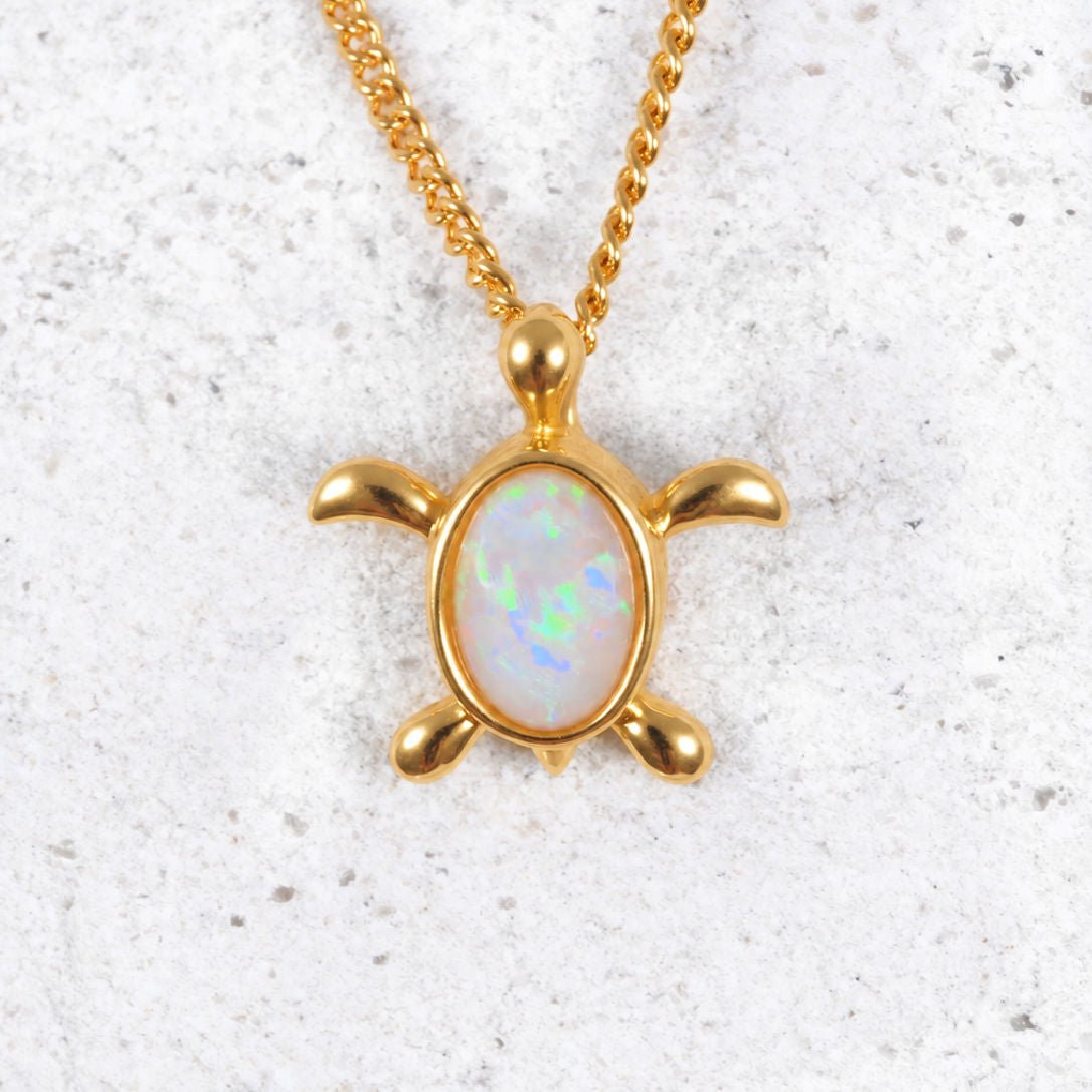 A gold plated silver turtle opal pendant featuring an iridescent white opal inlay on the shell, displayed on a textured light gray surface.